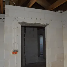 Surowy stan budynku w trakcie prac elektrycznych: widoczne kable prowadzone po ścianach z betonu komórkowego, zabezpieczone metalowymi uchwytami, puszki elektryczne w kolorze pomarańczowym...