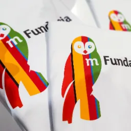 Białe koszulki z nadrukiem kolorowej sowy z logo 'm Fundacja' w zbliżeniu, lekko rozmyte tło.