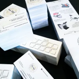 Stosy świeżo wydrukowanych materiałów reklamowych Body Space: wizytówki z logo, ulotki informacyjne o zabiegach modelowania sylwetki i karty lojalnościowe Super Slim z złotymi ramkami na czarnym tle.