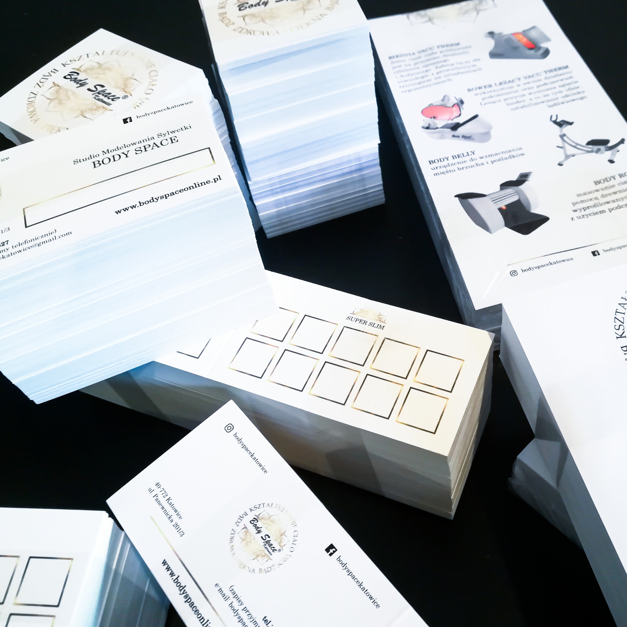 Stosy świeżo wydrukowanych materiałów reklamowych Body Space: wizytówki z logo, ulotki informacyjne o zabiegach modelowania sylwetki i karty lojalnościowe Super Slim z złotymi ramkami na czarnym tle.