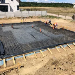 Przygotowanie zbrojenia fundamentu pod budowę domu jednorodzinnego, widoczna siatka zbrojeniowa, izolacja termiczna, rury instalacyjne, pracownicy przy pracy, betoniarka w tle.