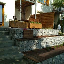 Marcin Pasieka - Ogród przydomowy z gabionami tworzącymi tarasy, drewniane skrzynie z kwiatami i drzewkami na górnym poziomie, schody z jasnego kamienia prowadzące do wejścia, drewniane panele ogrodzeniowe w tle.