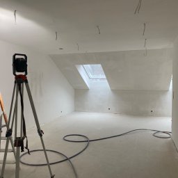 FLUGGER Gładzie Natryskowe i Malowanie - Poddasze po wyrównaniu ścian i sufitu, widoczne przewody elektryczne wystające z sufitu, drabina malarska, lampa na statywie i zwinięty wąż na podłodze.