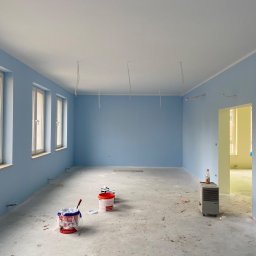 FLUGGER Gładzie Natryskowe i Malowanie - Pomalowane na błękitno wnętrze pokoju z trzema oknami, z widocznymi kablami zwisającymi z sufitu, z wiaderkami farby i grzejnikiem na podłodze przed otwartym przejściem do innego pomieszczenia.