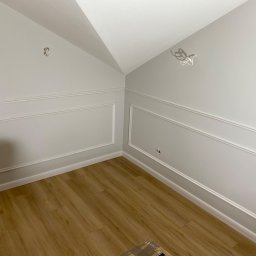 FLUGGER Gładzie Natryskowe i Malowanie - Pokój po remoncie z szarymi ścianami, sztukaterią i panelami podłogowymi, widoczne wystające kable elektryczne.