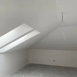 Poddasze po wyrównaniu ścian i sufitu, widoczne okno dachowe w skośnej ścianie, wystające przewody elektryczne i gładka, jasna powierzchnia ścian.
