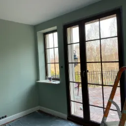 Świeżo pomalowane na pistacjowo ściany pokoju z dużymi oknami balkonowymi, zabezpieczona folią podłoga i drabina malarska w rogu, widoczne ślady taśmy malarskiej przy oknach.