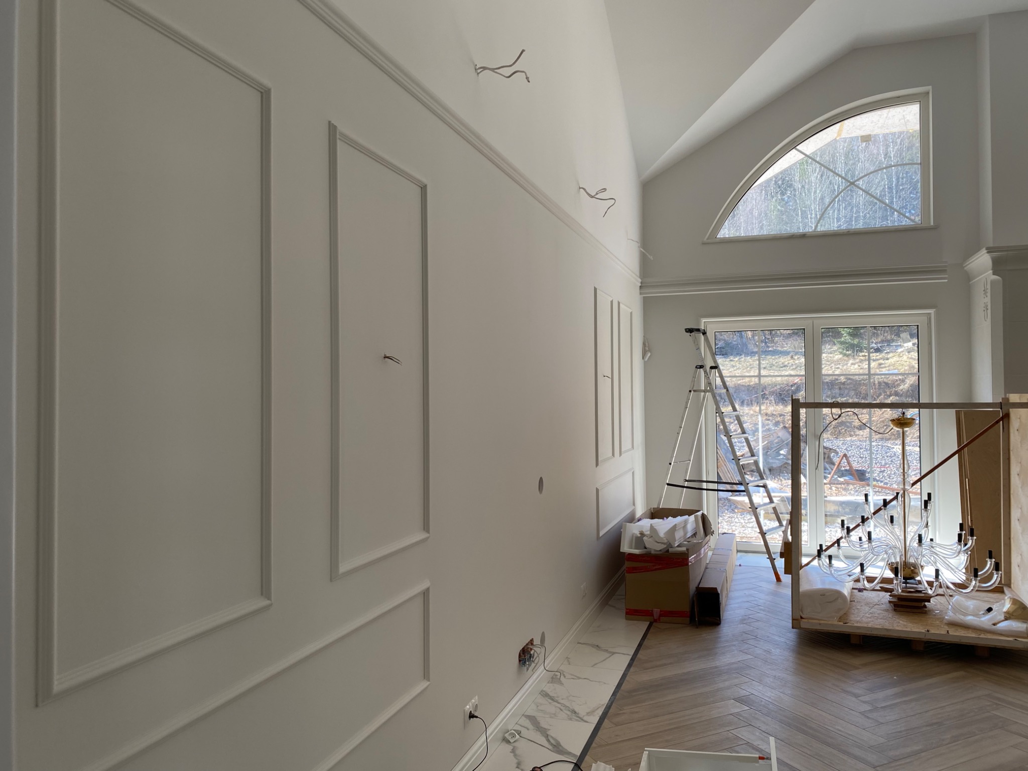 Ściana z dekoracyjnymi panelami, świeżo pomalowana na biało, z wystającymi przewodami elektrycznymi, widoczne przygotowania do montażu żyrandola.