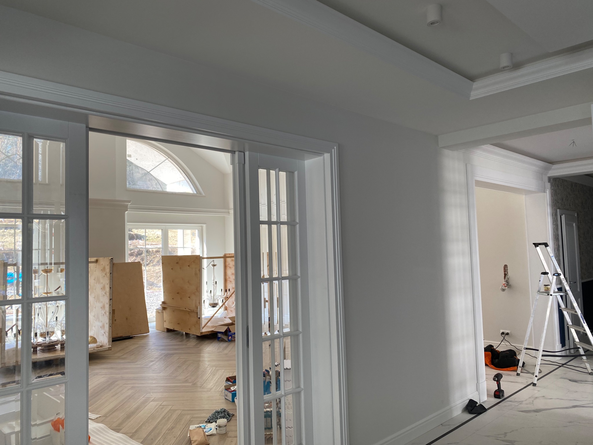 Wnętrze w trakcie prac wykończeniowych: białe ściany z listwami, drewniana podłoga w jodełkę, skrzynie z lampami widoczne przez przeszklone drzwi przesuwne, drabina oparta o ścianę w sąsiednim...