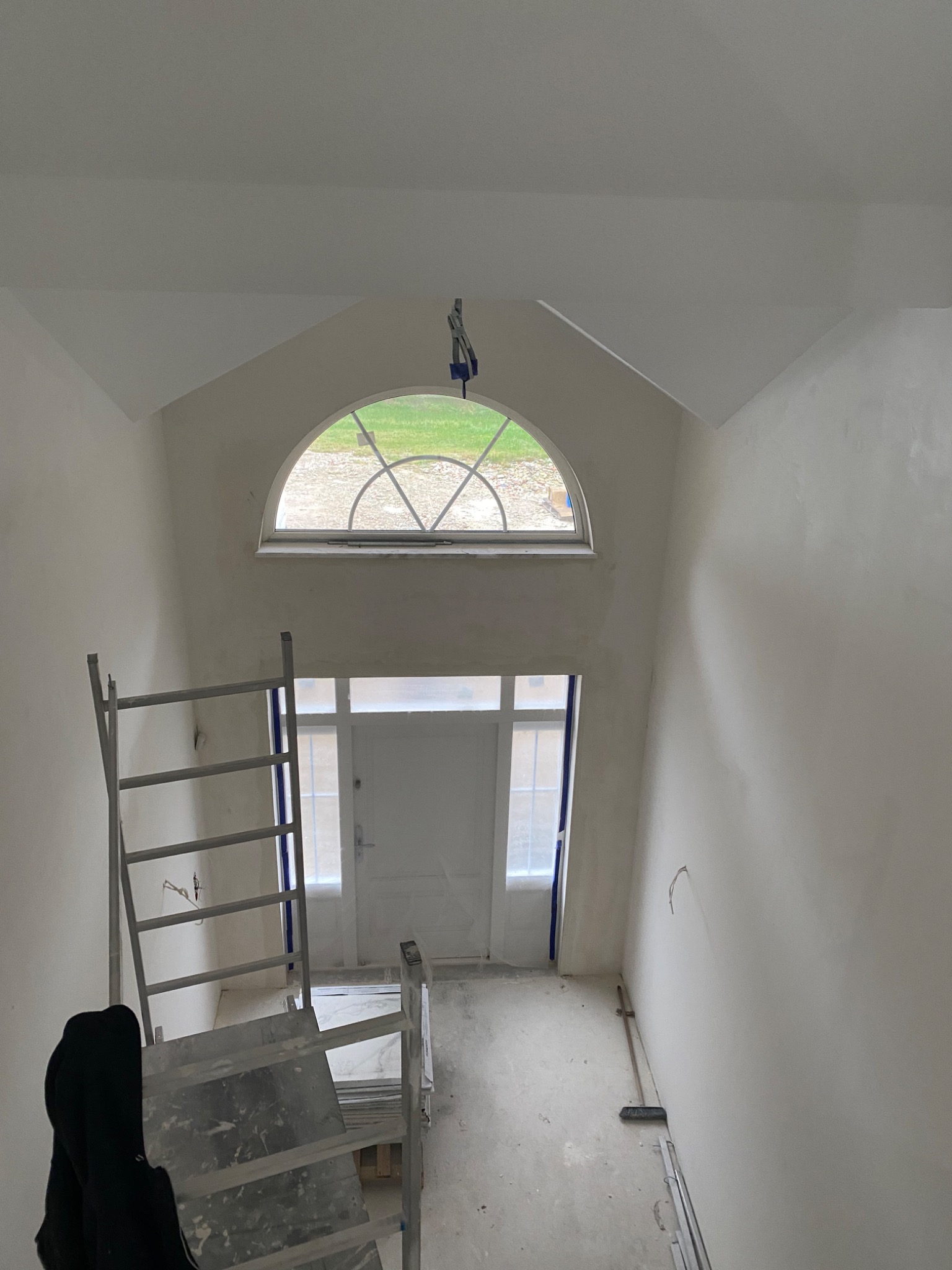 Klatka schodowa w trakcie remontu: ściany pokryte świeżą warstwą gładzi, widoczne rusztowanie i okno łukowe na górze. Na podłodze leżącą listwy przypodłogowe.