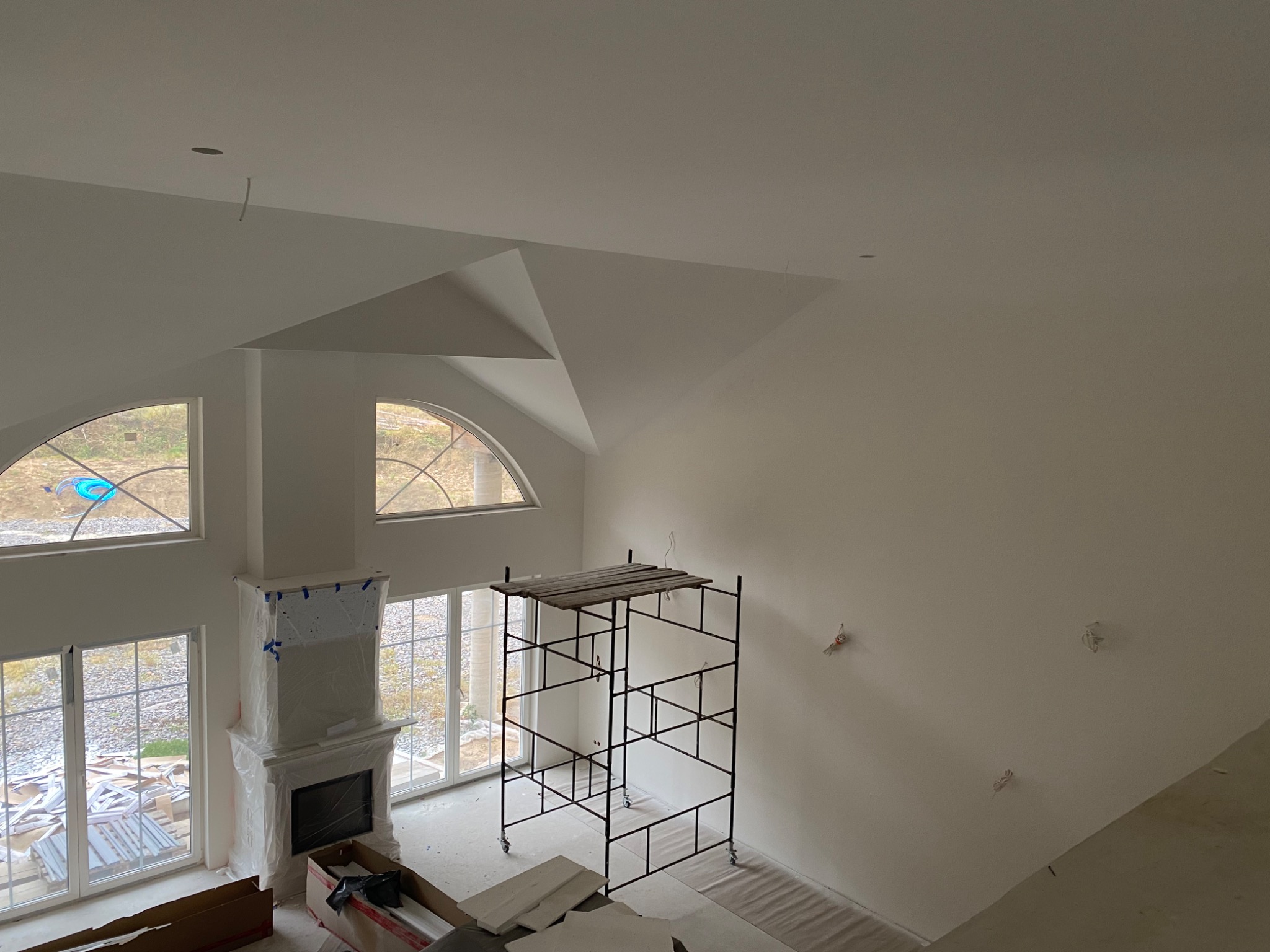 Wysokie pomieszczenie z gładkimi, białymi ścianami i sufitem, widoczne rusztowanie na kółkach, kominek zabezpieczony folią, duże okna z widokiem na zewnątrz, przygotowane materiały budowlane...