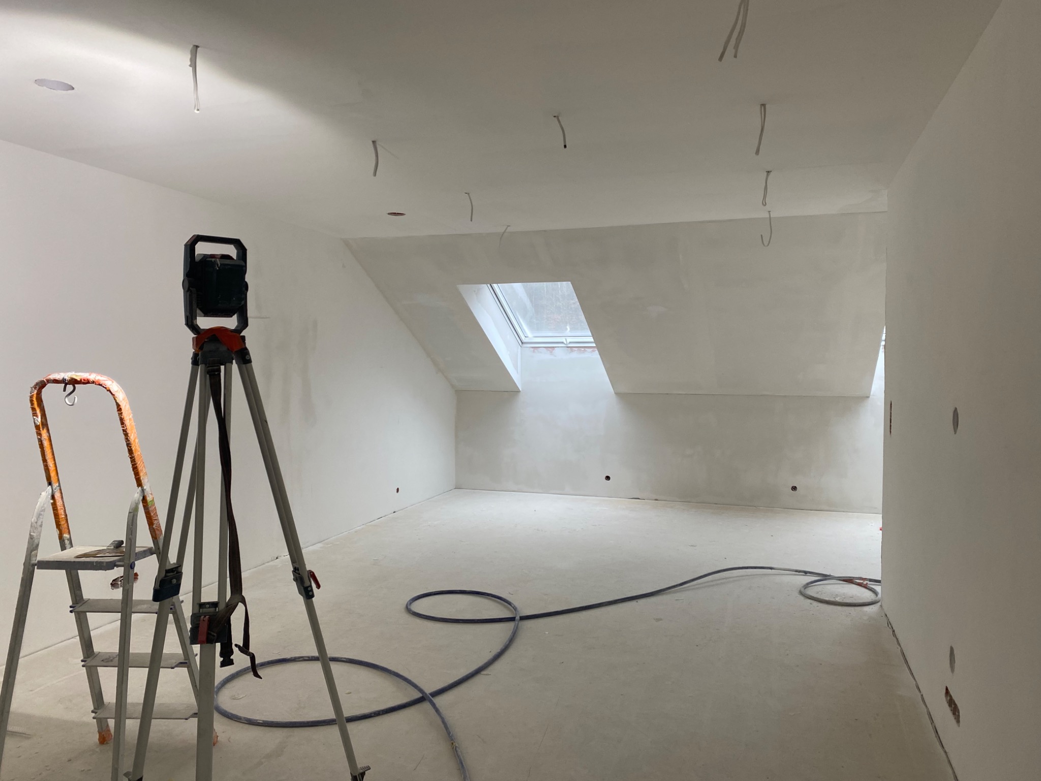 Poddasze po wyrównaniu ścian i sufitu, widoczne przewody elektryczne wystające z sufitu, drabina malarska, lampa na statywie i zwinięty wąż na podłodze.