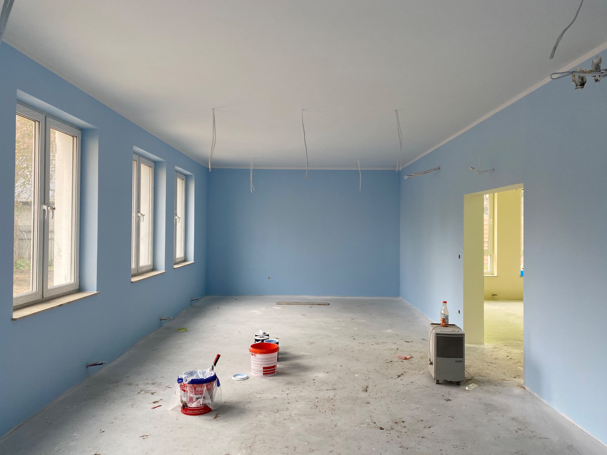 Pomalowane na błękitno wnętrze pokoju z trzema oknami, z widocznymi kablami zwisającymi z sufitu, z wiaderkami farby i grzejnikiem na podłodze przed otwartym przejściem do innego pomieszczenia.