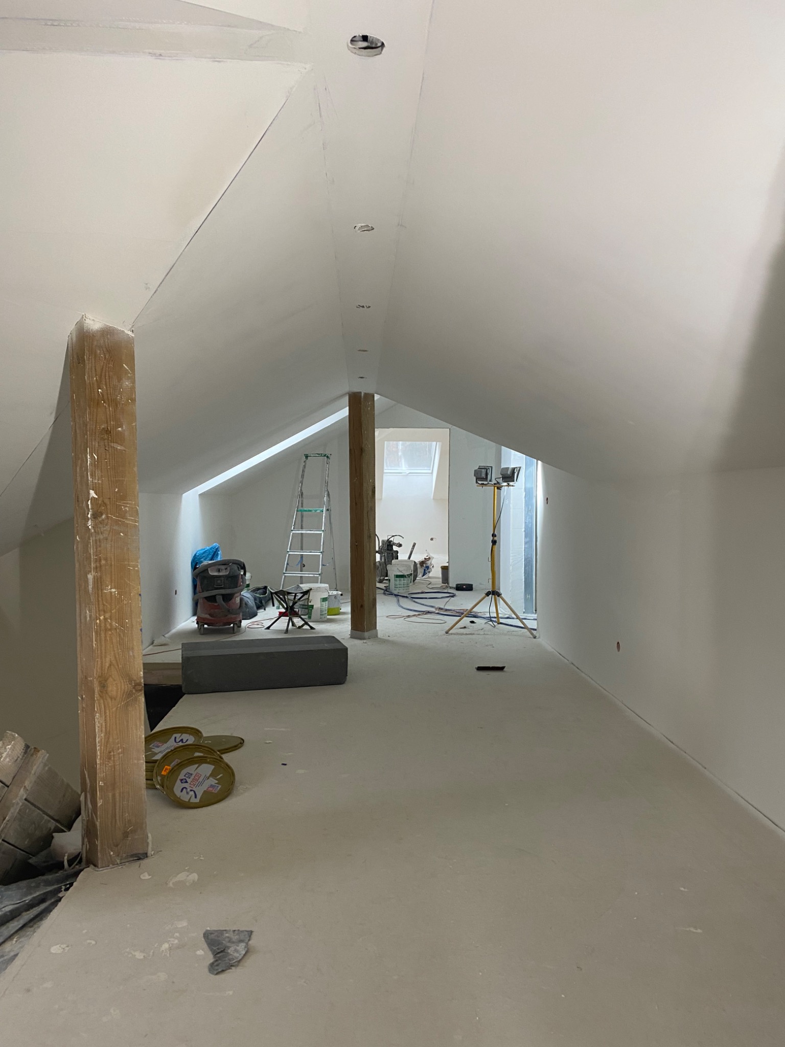 Poddasze w trakcie remontu: widoczne gładkie, białe ściany, drewniane słupy konstrukcyjne, drabina, lampa budowlana na trójnogu, odkurzacz przemysłowy i puste pojemniki po masie szpachlowej.