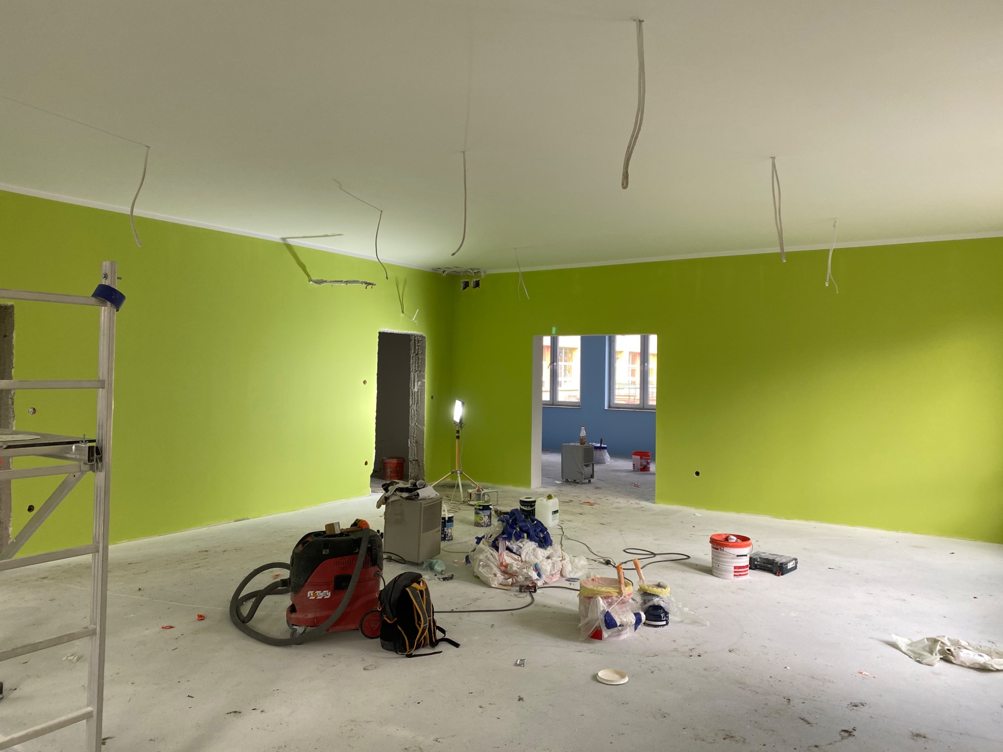Pokój w trakcie malowania na jaskrawy odcień zieleni, widoczny odkurzacz przemysłowy, drabina aluminiowa, kable zwisające z sufitu oraz akcesoria malarskie.