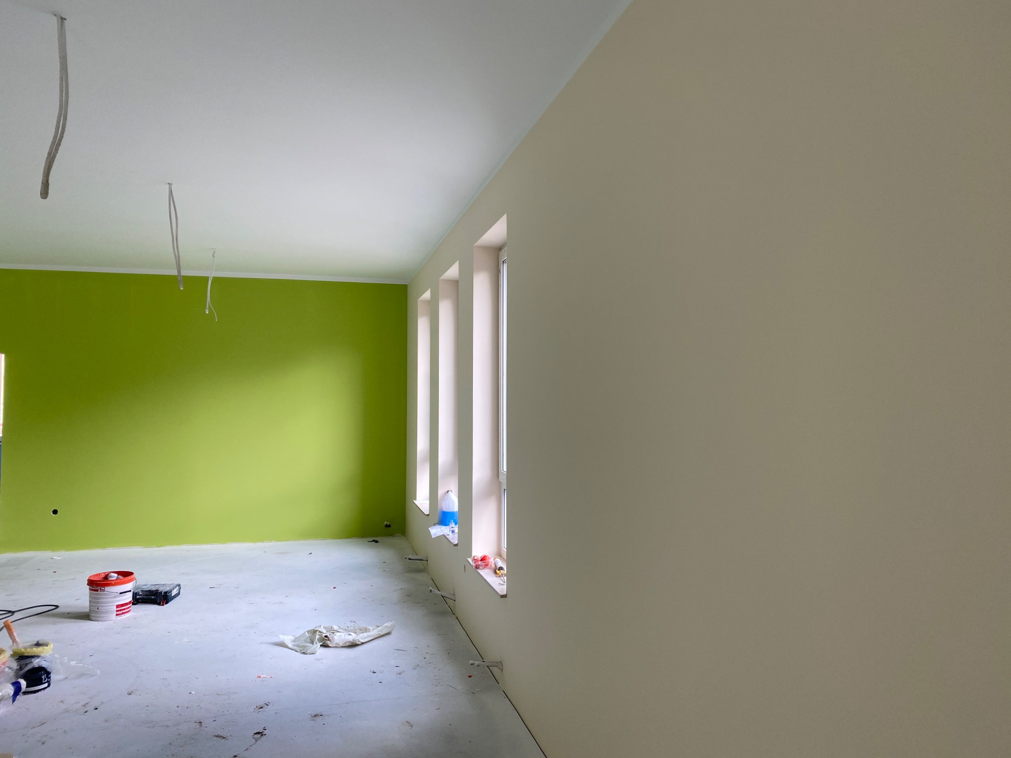 Wnętrze pokoju po częściowym pomalowaniu: jedna ściana zielona, pozostałe beżowe, widoczne puszki z farbą i akcesoria malarskie na podłodze.