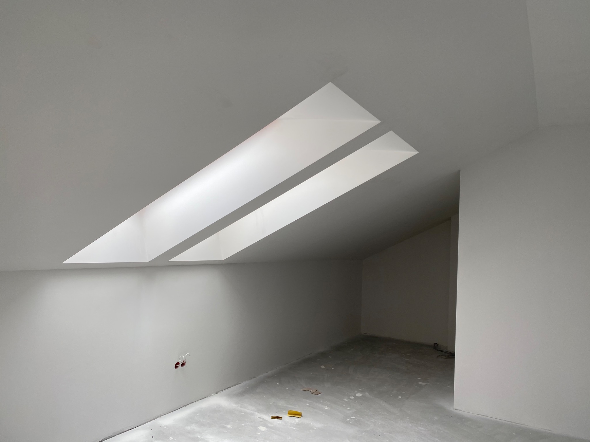 Poddasze po remoncie, ściany i sufit w jasnym odcieniu bieli, dwa okna dachowe w skośnym suficie, fragment podłogi z drobnymi zabrudzeniami, widoczne puszki elektryczne.