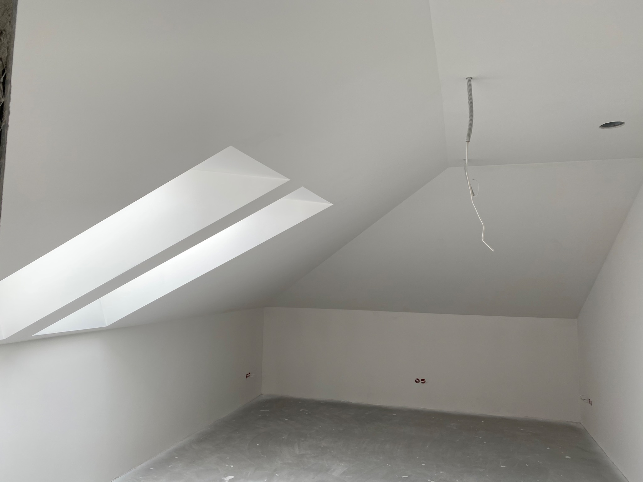 Poddasze po wyrównaniu ścian i sufitu, widoczne okno dachowe w skośnej ścianie, wystające przewody elektryczne i gładka, jasna powierzchnia ścian.