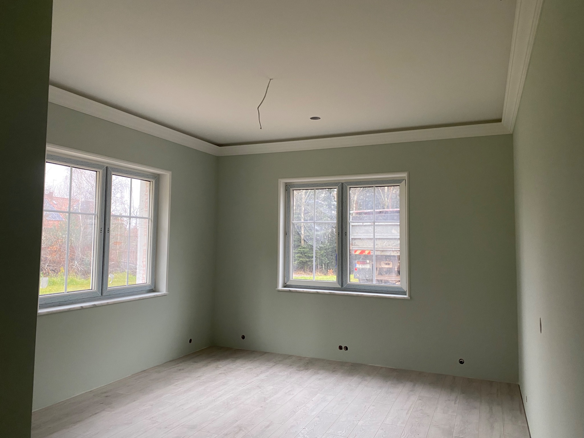 Pomalowane na jasnozielono wnętrze pokoju z dwoma oknami z widokiem na zewnątrz, widoczne otwory na gniazdka elektryczne w ścianach, jasna podłoga imitująca drewno, listwy dekoracyjne pod sufitem...