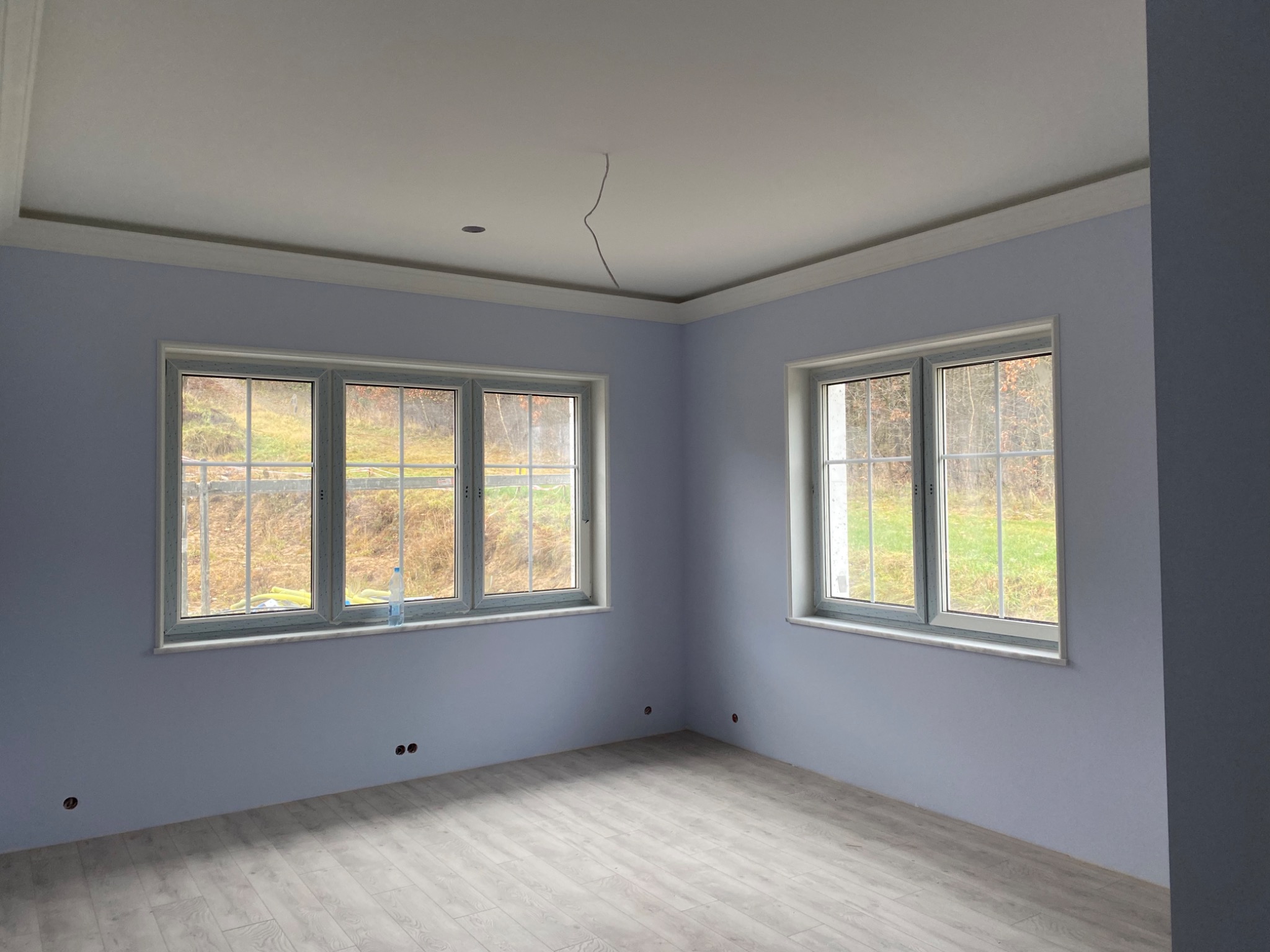 Pomalowane na jasny fiolet pokój z dwoma oknami, widoczne gniazdka elektryczne w ścianie, podłoga wyłożona panelami w odcieniach szarości, biały sufit z wystającym kablem.