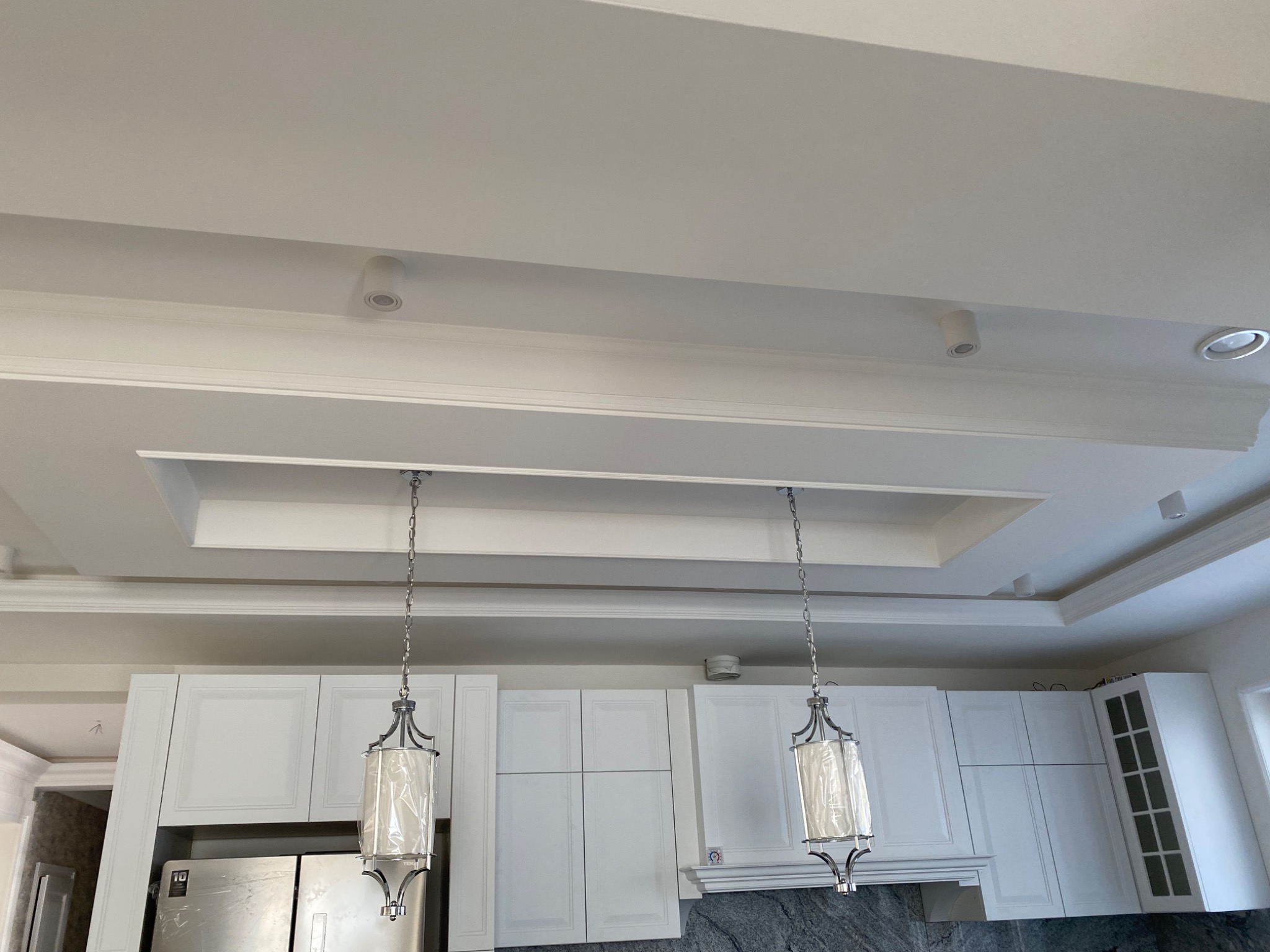 Wykończone wnętrze kuchni z białymi szafkami i ozdobnym sufitem podwieszanym z oświetleniem punktowym oraz dwoma wiszącymi lampami z łańcuchami, jeszcze w folii ochronnej.