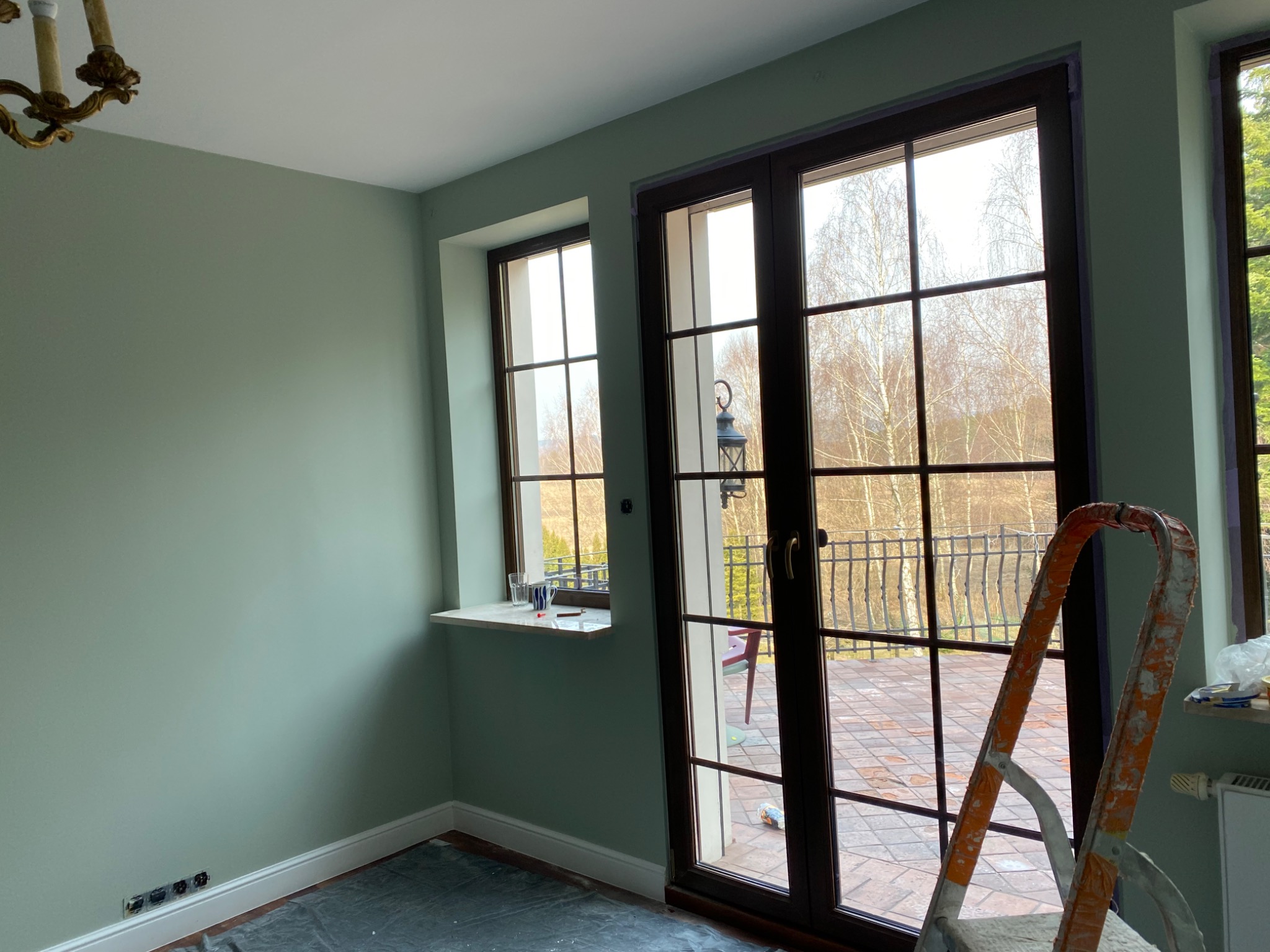 Świeżo pomalowane na pistacjowo ściany pokoju z dużymi oknami balkonowymi, zabezpieczona folią podłoga i drabina malarska w rogu, widoczne ślady taśmy malarskiej przy oknach.