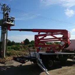 Pracownik w koszu podnośnika ISOLI PNT 185 przy słupie energetycznym, drugi pracownik przy skrzynce elektrycznej na ziemi, błękitne niebo w tle.