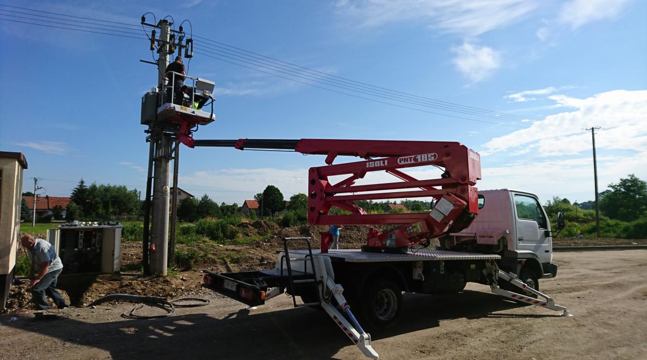 Pracownik w koszu podnośnika ISOLI PNT 185 przy słupie energetycznym, drugi pracownik przy skrzynce elektrycznej na ziemi, błękitne niebo w tle.
