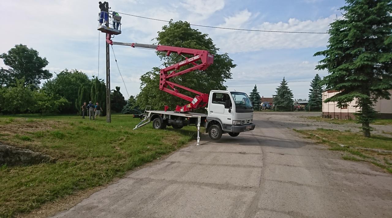 Biała ciężarówka z czerwonym ramieniem podnośnika koszowego ustawiona na betonowej nawierzchni, dwóch pracowników w koszu na wysokości słupa energetycznego, grupa osób obserwuje z dołu na trawniku.