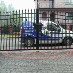 Czarna, metalowa brama wjazdowa z automatyką ramieniową, widoczny samochód serwisowy z naklejkami informacyjnymi firmy na tle domu.