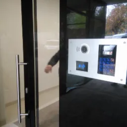 Nowoczesny panel zewnętrzny wideodomofonu z kamerą i czytnikiem kart zbliżeniowych, zamontowany na czarnej, szklanej fasadzie budynku, widoczny fragment drzwi wejściowych z metalowym uchwytem.