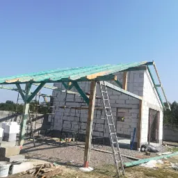 Wznoszona konstrukcja dachu z zielonymi elementami drewnianymi na budynku z białych bloczków, widoczna drabina i materiały budowlane na placu budowy.