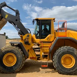 Żółta ładowarka Volvo z pełną łyżką materiału sypkiego nad skrzynią ładunkową ciężarówki, widoczne opony Michelin i operator w kabinie, błękitne niebo w tle.