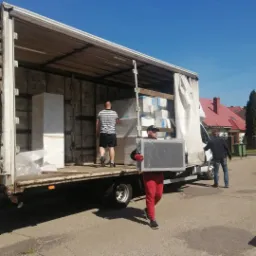 Mężczyzna w czerwonych spodniach i czapce niesie zapakowany w folię obraz na tle ciężarówki z załadunkiem mebli, drugi mężczyzna wchodzi do ciężarówki, a trzeci stoi obok.