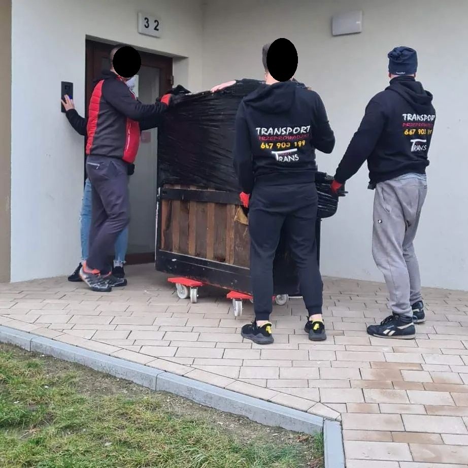 Czterech mężczyzn w odzieży roboczej transportuje mebel zabezpieczony czarną folią na wózku z kółkami przed budynkiem z numerem 32.