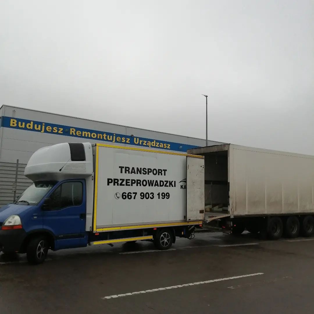 Niebiesko-biały samochód dostawczy z przyczepą, z napisem 'TRANSPORT PRZEPROWADZKI' i numerem telefonu, zaparkowany na parkingu przed budynkiem z napisem 'Budujesz Remontujesz Urządzasz', pochmurny...