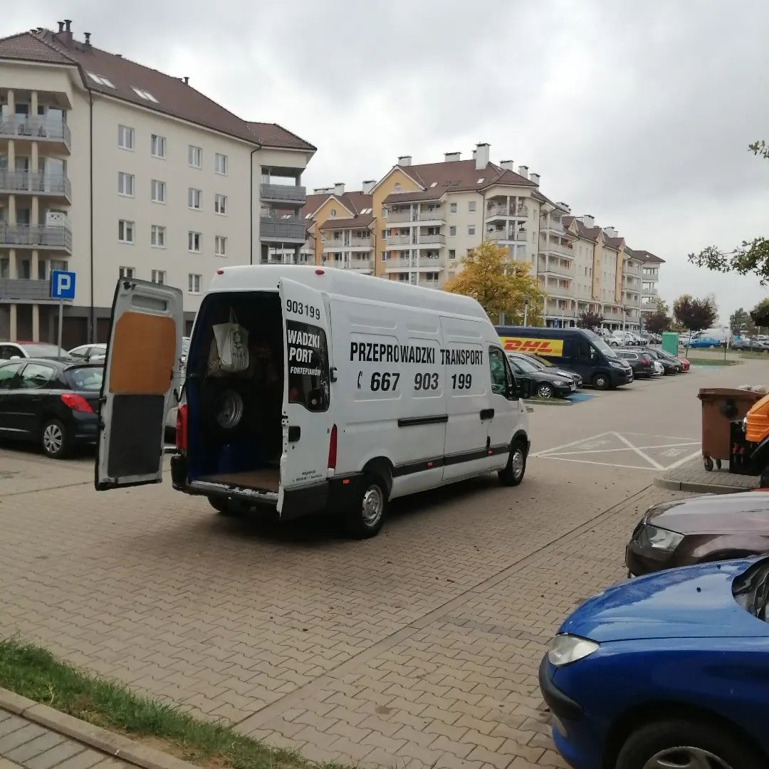 Biały bus z otwartymi drzwiami bagażnika na parkingu, widoczne wnętrze z workami i oponą, na karoserii napis 'Przeprowadzki Transport' i numer telefonu.