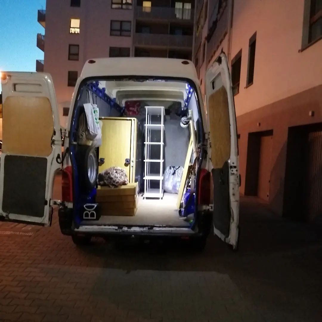 Otwarty tył białego busa zaparkowanego przed budynkiem mieszkalnym, załadowany meblami: drzwi, regał, skrzynia, koło zapasowe i torby.