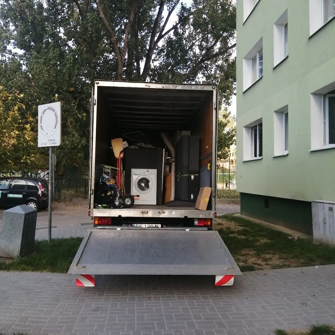 Otwarta paka ciężarówki załadowana sprzętem AGD i meblami podczas dnia, widoczna pralka, karton, miotła i inne przedmioty, tło stanowi blok mieszkalny i drzewa.