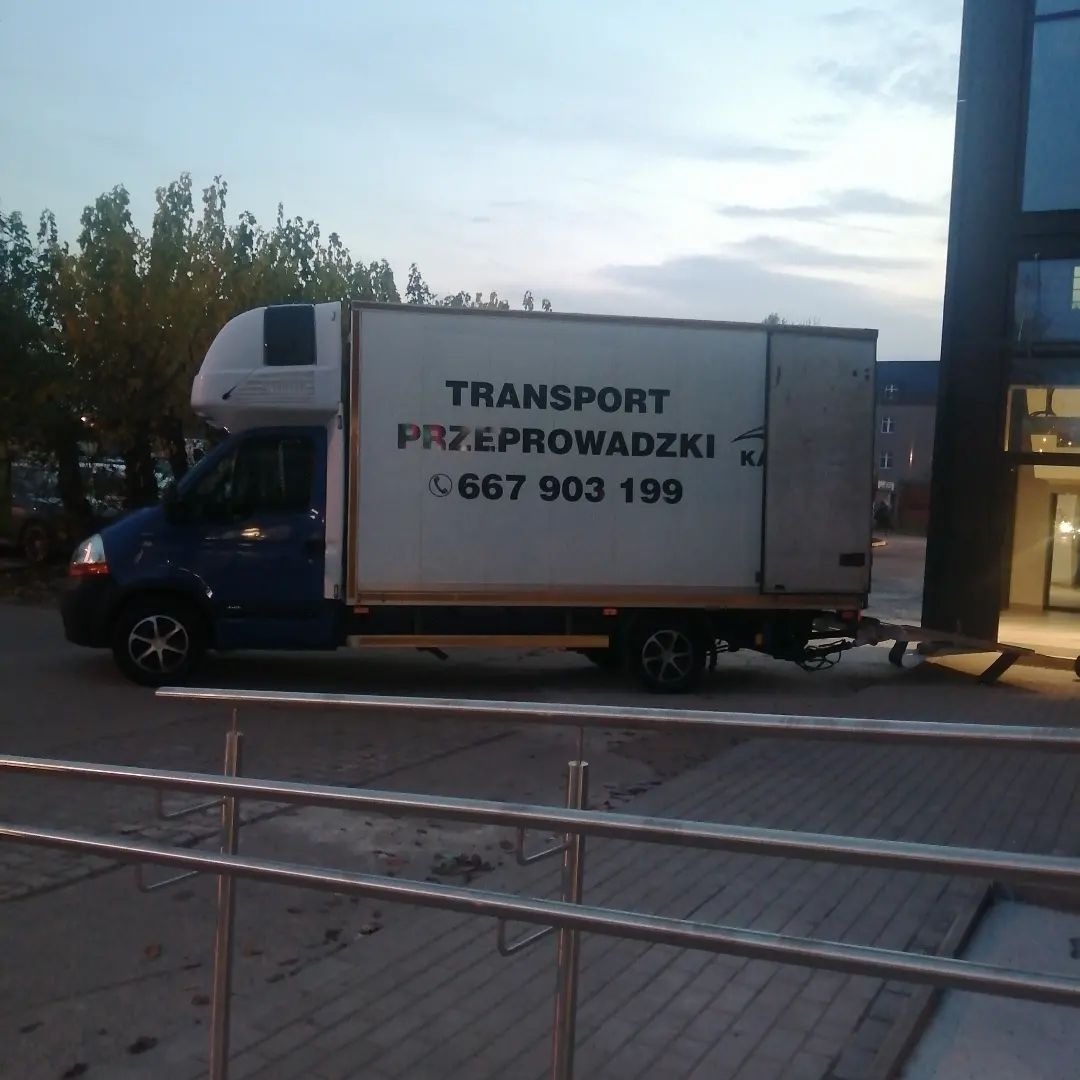 Niebieski bus transportowy z białą nadbudówką i napisem 'TRANSPORT PRZEPROWADZKI 667 903 199' zaparkowany przy nowoczesnym budynku z metalowymi barierkami.