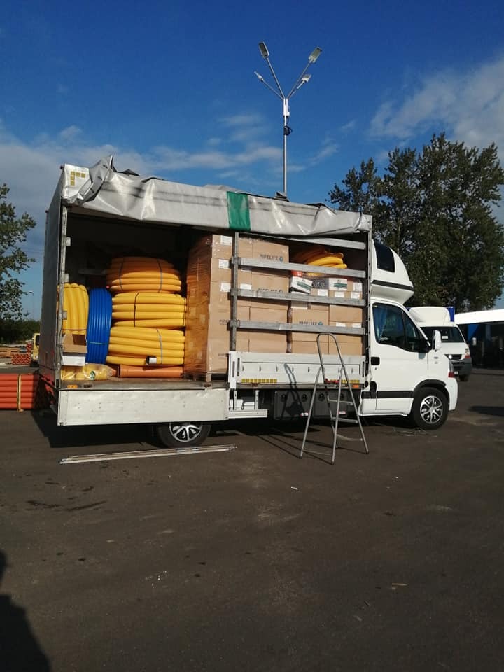 Otwarta paka białego samochodu dostawczego wypełniona kartonami z napisem PIPELIFE i zwiniętymi rurami w kolorach żółtym i niebieskim, drabina oparta o bok, na zewnątrz plac z latarnią.