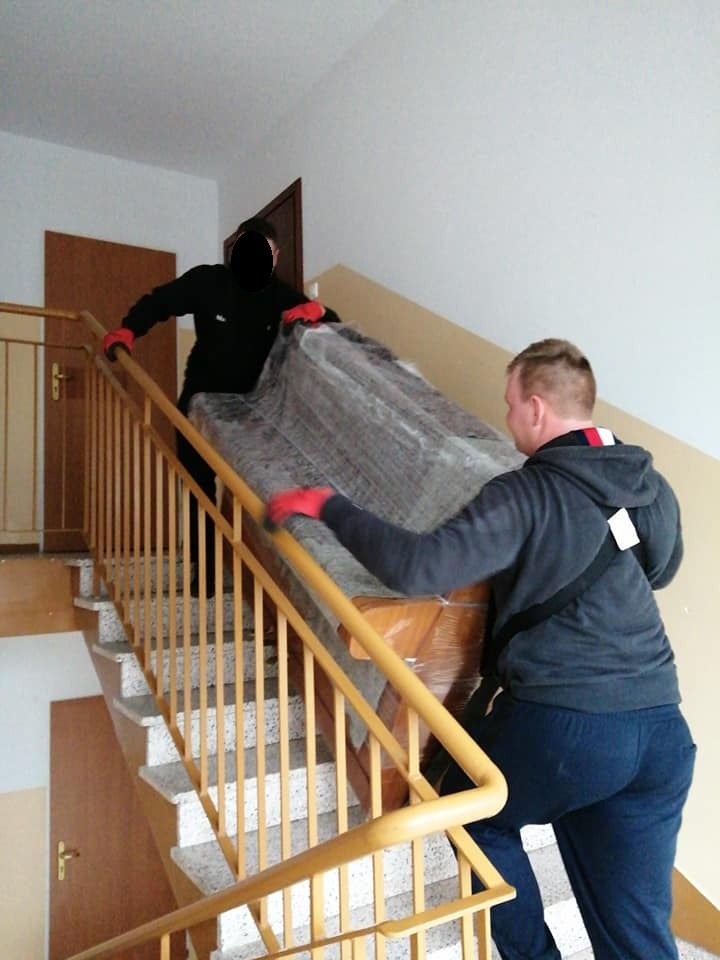 Dwóch mężczyzn w czerwonych rękawicach wnosi po schodach zabezpieczony folią fortepian, jeden z nich opiera się o żółtą barierkę.