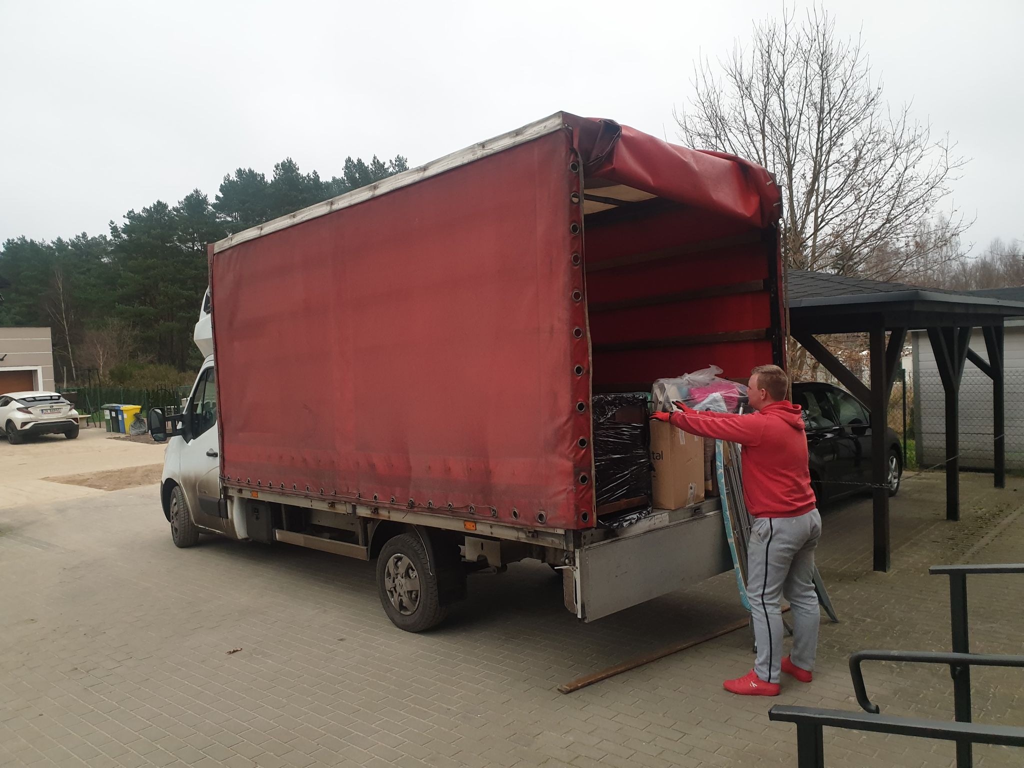 Mężczyzna w czerwonej bluzie i czerwonych butach wkłada do otwartej ciężarówki z czerwoną plandeką zapakowane meble i kartony.