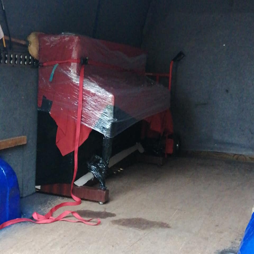 Pianino zabezpieczone folią i czerwonym materiałem, umieszczone wewnątrz ciemnej przestrzeni ładunkowej, prawdopodobnie podczas transportu.