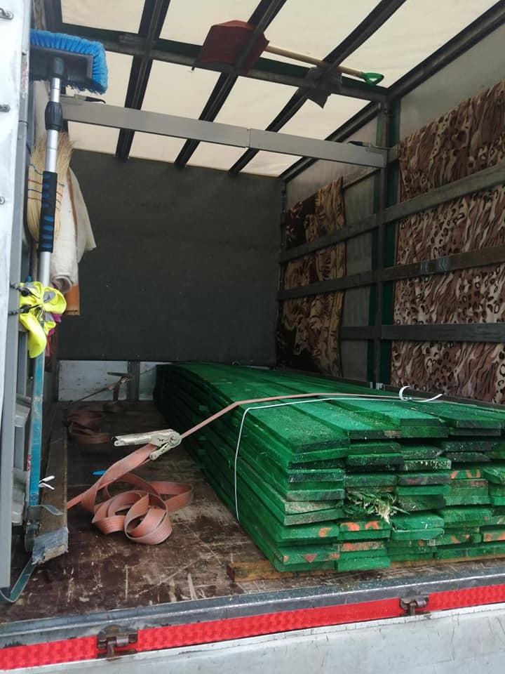 Wnętrze samochodu dostawczego z zabezpieczonym stosem pomalowanych na zielono desek, widoczna także miotła, szufelka i koce ochronne.