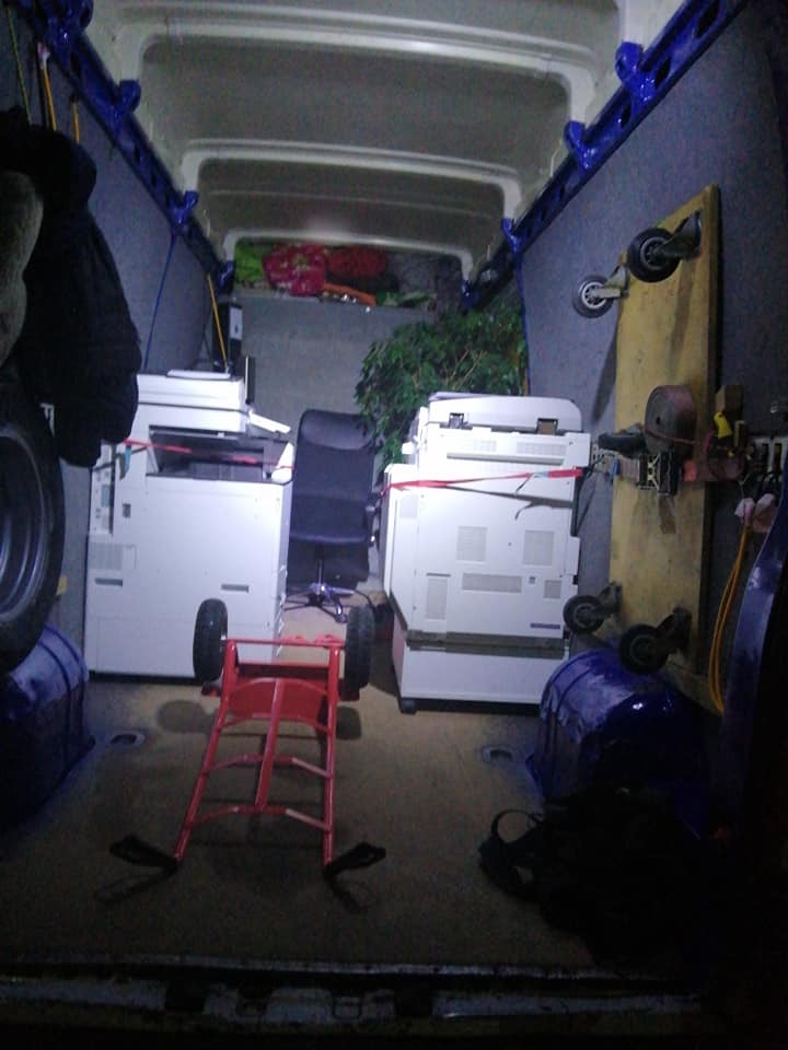 Wnętrze furgonetki z dwoma zabezpieczonymi kserokopiarkami, biurowym krzesłem i rośliną doniczkową, przygotowane do transportu, widoczny wózek transportowy.