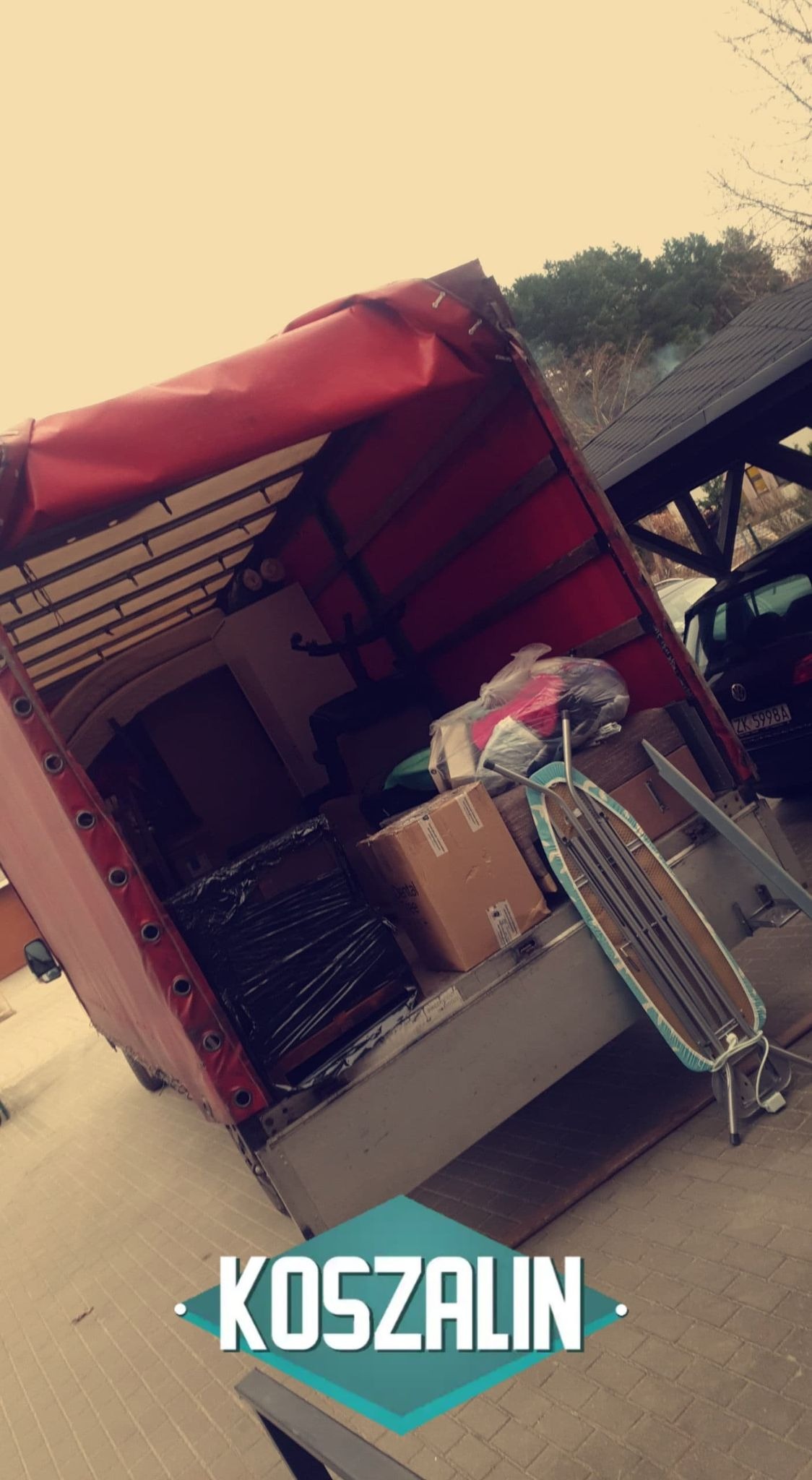 Wnętrze otwartej ciężarówki z czerwonym plandekowym dachem załadowanej meblami, kartonami i deską do prasowania, na tle kostki brukowej i zaparkowanego samochodu osobowego, z napisem 'Koszalin'...
