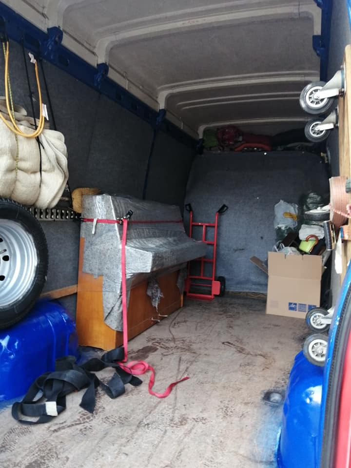 Wnętrze busa z zabezpieczonym pianinem, wózkiem transportowym, zapasowym kołem i workami, przygotowane do transportu.