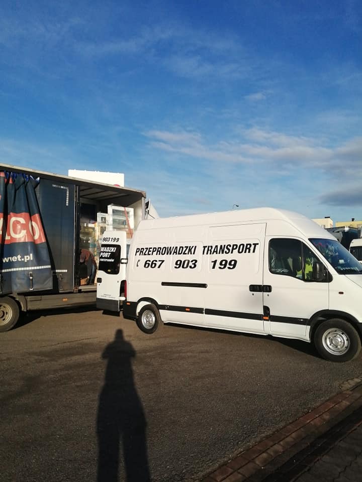 Biały bus z napisem 'Przeprowadzki Transport 667 903 199' na tle załadowanej ciężarówki i błękitnego nieba, cień fotografa w dolnej części kadru.