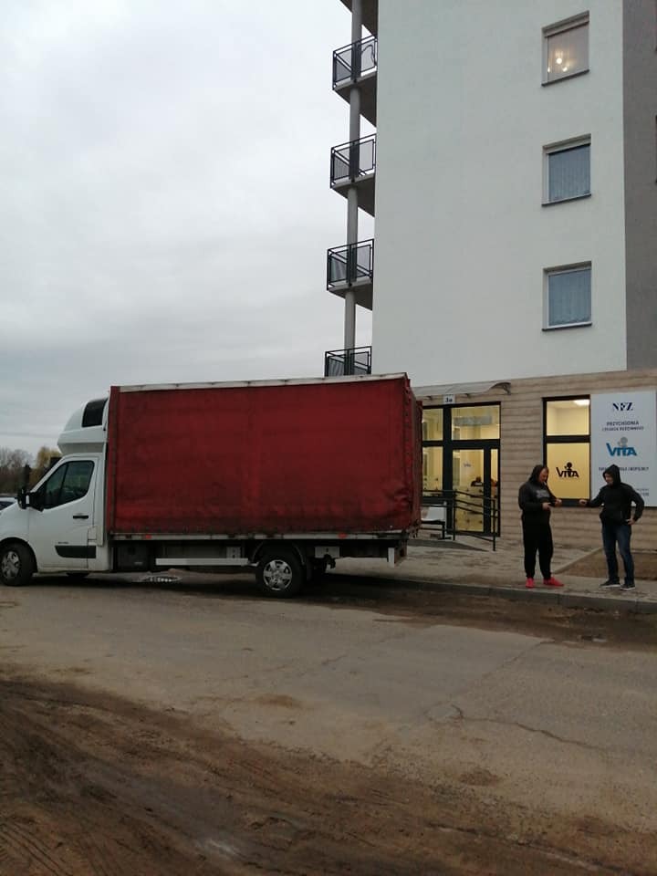 Czerwony samochód dostawczy zaparkowany przed nowoczesnym budynkiem z balkonami, dwóch mężczyzn rozmawia obok wejścia.